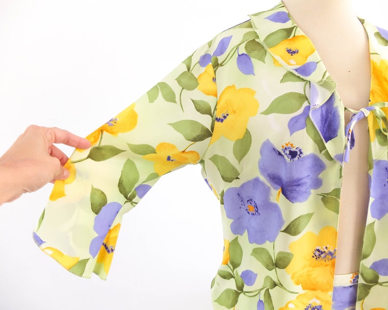 May include: A light green, yellow, and purple floral print blouse with a ruffled collar and a tie at the neck.