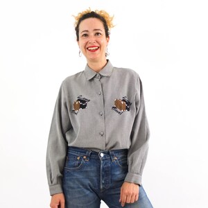 May include: A gray button-down shirt with long sleeves and a collar. The shirt has an embroidered design on the front that says "Long Travel" with a suitcase and a plane. The shirt is worn by a person with blonde hair and a smile.