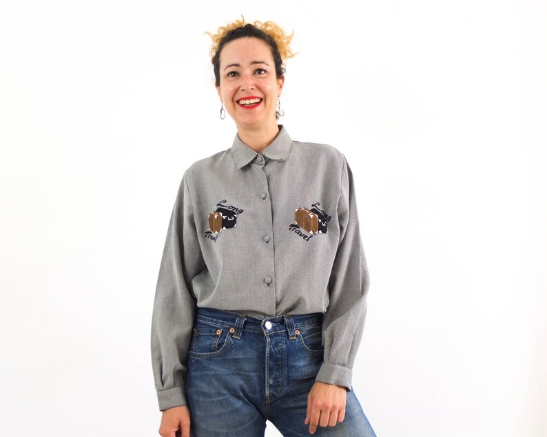 May include: A gray button-down shirt with long sleeves and a collar. The shirt has an embroidered design on the front that says "Long Travel" with a suitcase and a plane. The shirt is worn by a person with blonde hair and a smile.