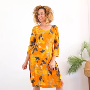 Op de afbeelding: Een knielange jurk in een levendige oranje tint met een donkerblauw en lichtbruin bladpatroon. Het kledingstuk heeft driekwartmouwen, een ronde halslijn en zijsplitten die vanaf de zoom omhoog lopen.
