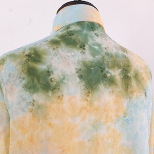 Op de afbeelding: Een tie-dye shirt in geel, groen en blauw. Het shirt heeft een opstaande kraag en delicate bloemenborduursels. De lichte stof suggereert een ontwerp voor warmer weer. Een uniek kledingstuk.
