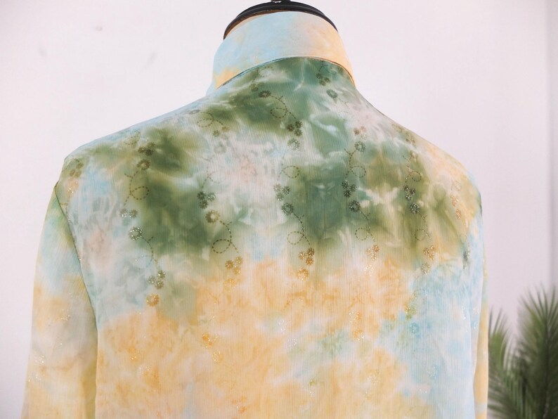 Op de afbeelding: Een tie-dye shirt in geel, groen en blauw. Het shirt heeft een opstaande kraag en delicate bloemenborduursels. De lichte stof suggereert een ontwerp voor warmer weer. Een uniek kledingstuk.