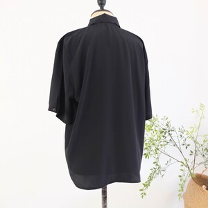 May include: A black short-sleeved button-down shirt displayed on a mannequin. The shirt has a collar and a loose fit. The fabric appears to be lightweight and flowing. The background is a plain white wall with a touch of greenery.