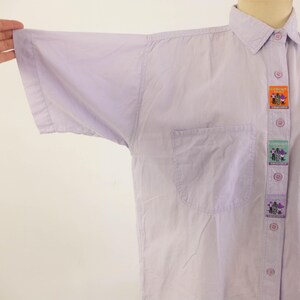 May include: A light purple button-down shirt with short sleeves and a chest pocket. The shirt features decorative patches down the button line. The patches have colorful illustrations. The shirt is made of a lightweight fabric.