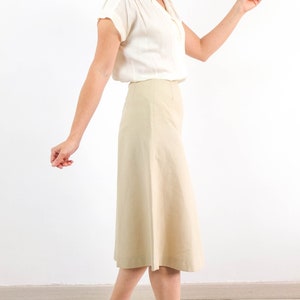 May include: A woman wearing a white blouse and a beige skirt. The skirt is knee-length and has a slight A-line shape.