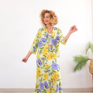 May include: A woman wearing a green and yellow floral print jacket and skirt set. The jacket has a light blue floral pattern and a white collar. The skirt is a midi length and has a light blue and yellow floral pattern.