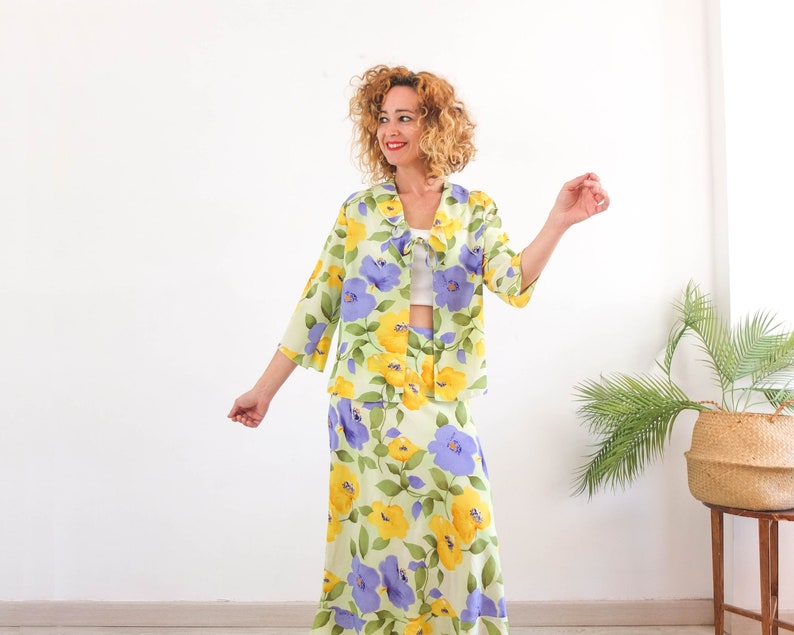 May include: A woman wearing a green and yellow floral print jacket and skirt set. The jacket has a light blue floral pattern and a white collar. The skirt is a midi length and has a light blue and yellow floral pattern.