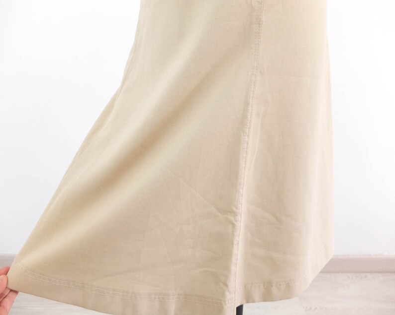 May include: A beige skirt with a straight cut and a slight flare at the bottom. The skirt has a seam down the back and a hemmed edge.