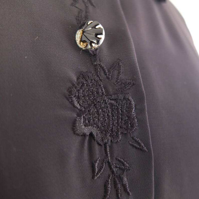 May include: Close-up of a dark gray garment with intricate black floral embroidery. A decorative button with a black shell design and gold accents is visible. The fabric has a smooth texture.