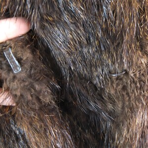 May include: Close-up of a brown fur coat with a metal hook and loop closure. The fur is soft and fluffy.