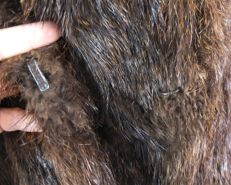 May include: Close-up of a brown fur coat with a metal hook and loop closure. The fur is soft and fluffy.