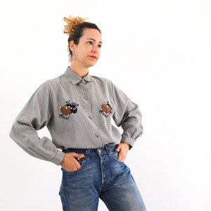 May include: A gray long-sleeved button-up shirt with a relaxed fit. The shirt features two embroidered brown suitcases with the text "Long Travel" on each.