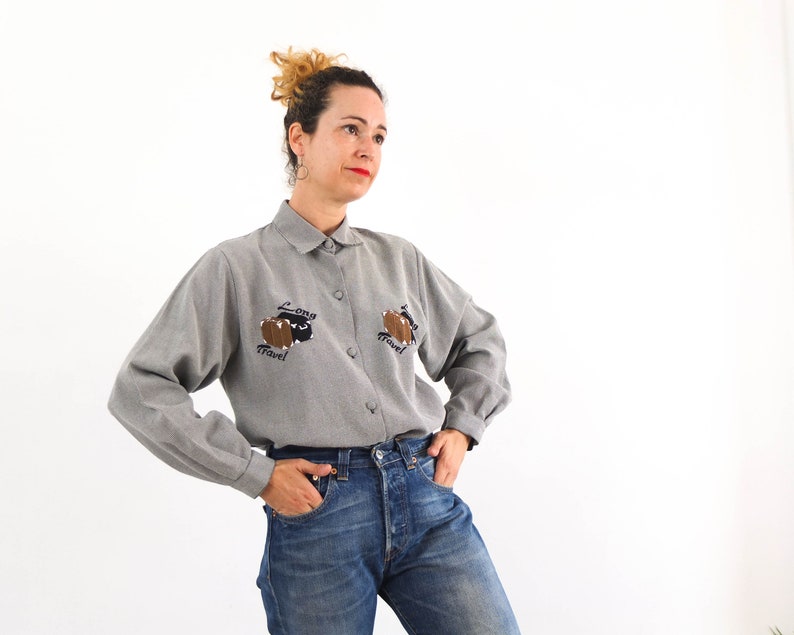 May include: A gray long-sleeved button-up shirt with a relaxed fit. The shirt features two embroidered brown suitcases with the text "Long Travel" on each.
