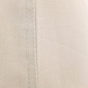 May include: Close-up of a beige fabric with a single seam running down the centre. The fabric has a subtle texture and a slightly off-white colour.