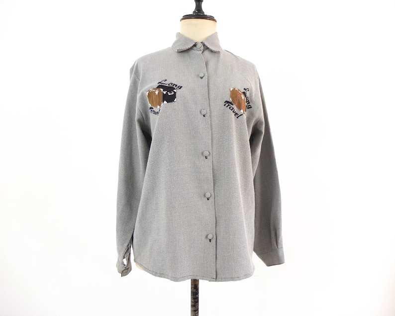 May include: A gray and white checkered button-up shirt with long sleeves and a collared neckline. The shirt features embroidered brown luggage with the text "Long Travel" on the left and right sides.