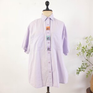 May include: A light purple button-down shirt with short sleeves, displayed on a mannequin. The shirt features a series of decorative patches down the front, each with a colorful design. The shirt has two front pockets and a classic collar.