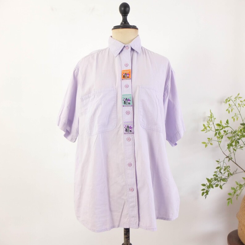 May include: A light purple button-down shirt with short sleeves, displayed on a mannequin. The shirt features a series of decorative patches down the front, each with a colorful design. The shirt has two front pockets and a classic collar.