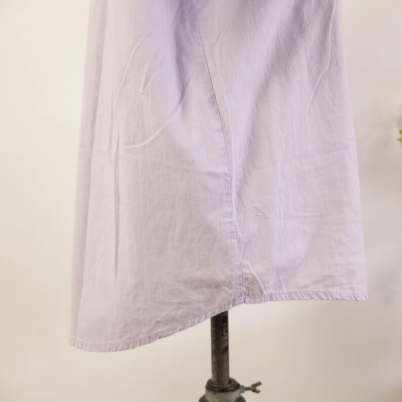 May include: A light purple dress on a metal stand. The dress is made of a soft, lightweight fabric and has a simple, flowing design. The hemline is slightly uneven, adding to the dress's casual appeal. The dress is likely vintage or handmade.