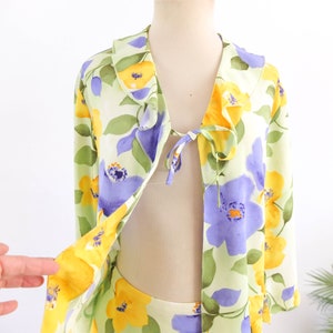 May include: A light green, yellow, and purple floral print top with a ruffled collar and a tie at the neckline.