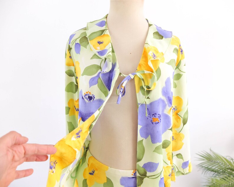 May include: A light green, yellow, and purple floral print top with a ruffled collar and a tie at the neckline.