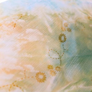 Op de afbeelding: Close-up van een transparante stof met een aquareleffect in tinten blauw, geel en groen. Delicate gouden bloemenborduurwerk accentueert de stof en cre&euml;ert een zachte, vloeiende esthetiek. Ideaal voor sjaals of lichte kleding.