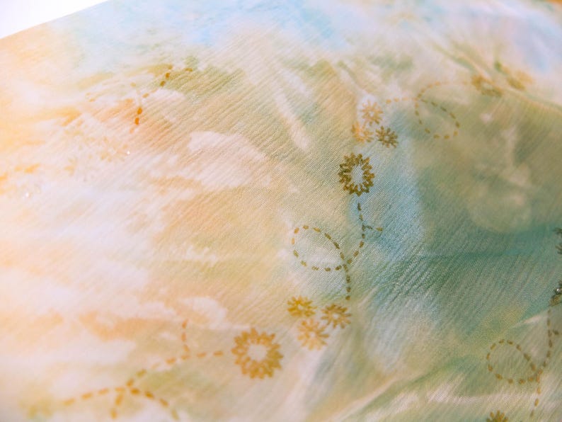 Op de afbeelding: Close-up van een transparante stof met een aquareleffect in tinten blauw, geel en groen. Delicate gouden bloemenborduurwerk accentueert de stof en cre&euml;ert een zachte, vloeiende esthetiek. Ideaal voor sjaals of lichte kleding.
