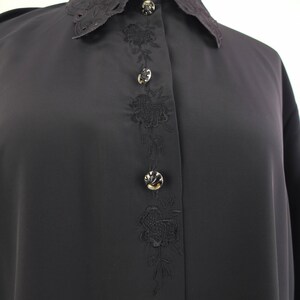 May include: A black blouse with a lace-trimmed collar and floral embroidery down the front. The blouse features decorative buttons and long sleeves. The fabric appears to be a smooth, lightweight material.