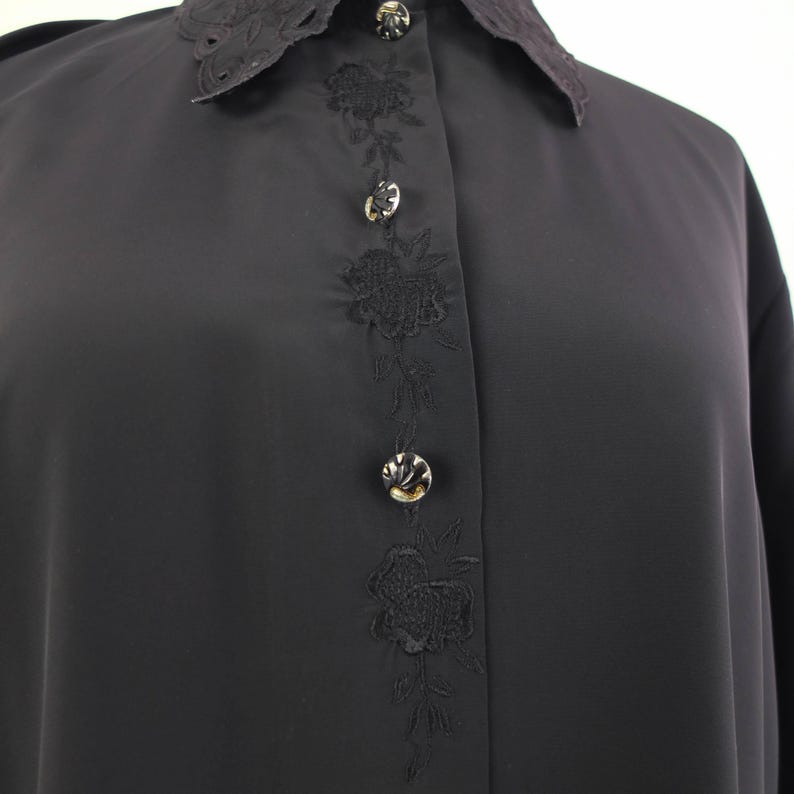 May include: A black blouse with a lace-trimmed collar and floral embroidery down the front. The blouse features decorative buttons and long sleeves. The fabric appears to be a smooth, lightweight material.