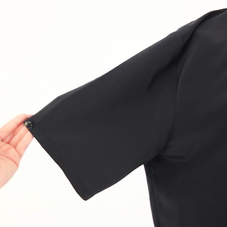 May include: Close-up of a black short-sleeved garment. The sleeve is visible, with a button detail at the cuff. The fabric appears smooth and opaque, with a slight sheen. The garment is against a white background.