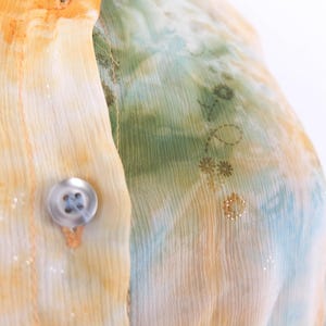 Op de afbeelding: Close-up van een doorschijnende blouse met knoopsluiting. De stof heeft een aquareleffect in geel, oranje, groen en blauw, met een delicaat bloemmotief. Een doorzichtige knoop is zichtbaar.