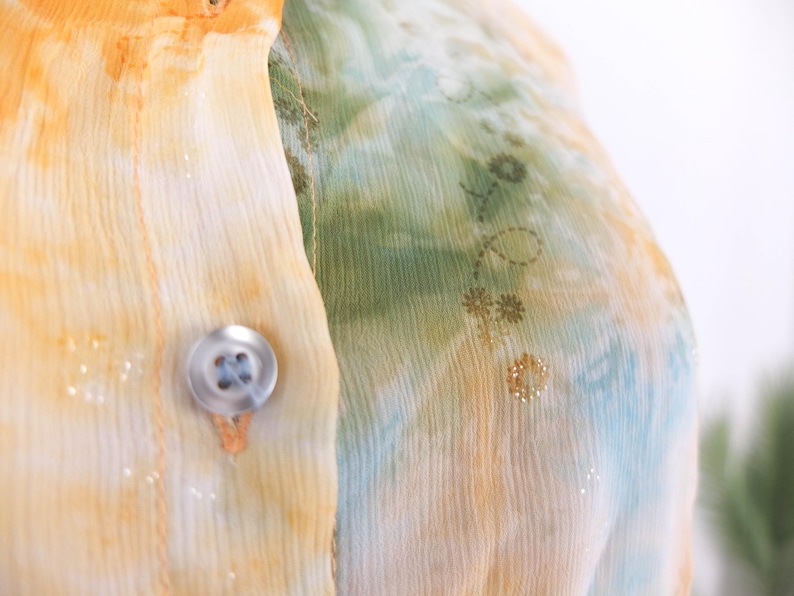 Op de afbeelding: Close-up van een doorschijnende blouse met knoopsluiting. De stof heeft een aquareleffect in geel, oranje, groen en blauw, met een delicaat bloemmotief. Een doorzichtige knoop is zichtbaar.