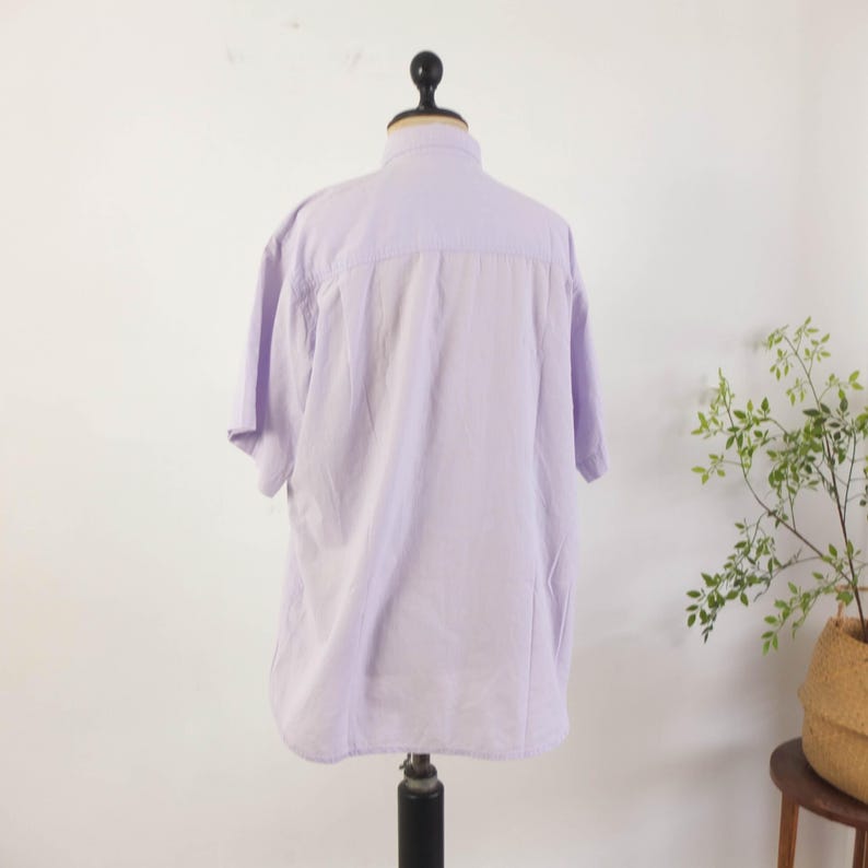 May include: A light purple short-sleeved button-down shirt displayed on a mannequin. The shirt has a classic collar and a relaxed fit. The back of the shirt is visible, showing the yoke and the overall shape of the garment. The shirt is made of a lightweight fabric.