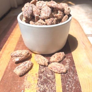 May include: A white ceramic bowl filled with brown, sugar-coated pecans. Several individual sugar-coated pecans are scattered around the base of the bowl. The bowl sits on a wooden board featuring alternating light and dark wood grain stripes.