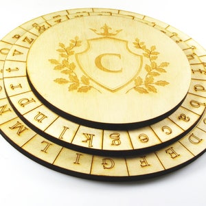 Può includere: Un puzzle circolare in legno con tre strati rotanti. Ogni strato ha una sequenza alfabetica diversa. Lo strato superiore presenta uno scudo con una corona e la lettera "C" al suo interno.