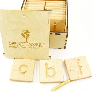 K&ouml;nnte beinhalten: Holzkiste mit einem Deckel, auf dem "MONTESSORI Lernen durch Entdecken" steht, und darin enthalten sind Holzkacheln mit Buchstaben darauf. Die Kacheln sind in einem Raster angeordnet und daneben liegt ein kleiner Holzstift.