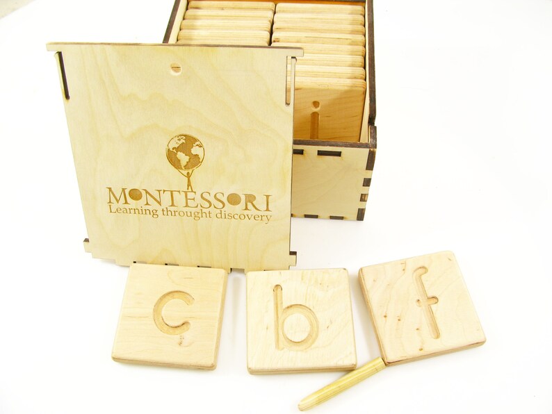 K&ouml;nnte beinhalten: Holzkiste mit einem Deckel, auf dem "MONTESSORI Lernen durch Entdecken" steht, und darin enthalten sind Holzkacheln mit Buchstaben darauf. Die Kacheln sind in einem Raster angeordnet und daneben liegt ein kleiner Holzstift.