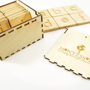 K&ouml;nnte beinhalten: Holzkiste mit einem Satz Alphabet-Kacheln und einem kleinen Holzstift. Die Kiste ist mit den Worten "Montessori Lernen durch Entdecken" graviert.