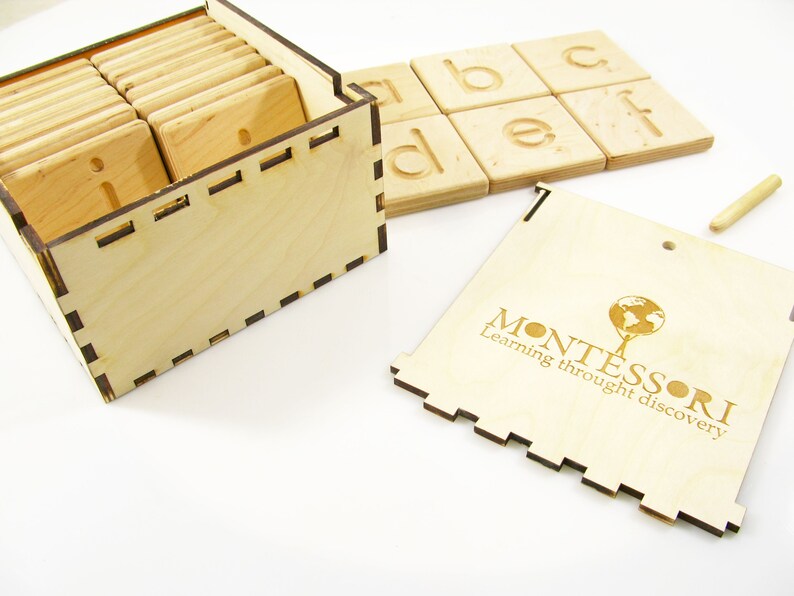 K&ouml;nnte beinhalten: Holzkiste mit einem Satz Alphabet-Kacheln und einem kleinen Holzstift. Die Kiste ist mit den Worten "Montessori Lernen durch Entdecken" graviert.