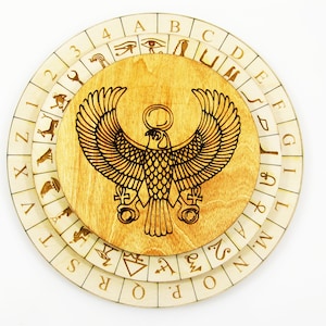 Könnte beinhalten: Ein rundes Holz-Divinationsbrett mit drei drehbaren Ringen. Der mittlere Ring zeigt ein eingraviertes Bild eines geflügelten ägyptischen Gottes mit einer Sonnenscheibe über seinem Kopf. Die äußeren Ringe sind mit Buchstaben und Zahlen beschriftet.