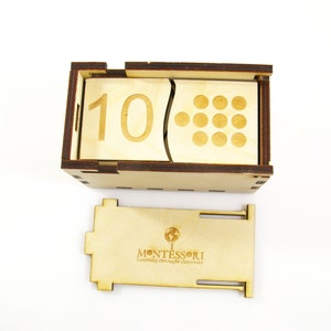 May include: Wooden box with a sliding lid, containing a number block with the number 10 and a corresponding number of circles. The box also has a separate wooden piece with the word "MONTESSORI" and the phrase "Learning through discovery".