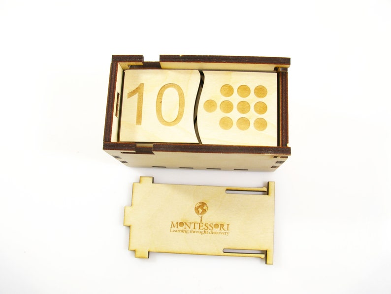 May include: Wooden box with a sliding lid, containing a number block with the number 10 and a corresponding number of circles. The box also has a separate wooden piece with the word "MONTESSORI" and the phrase "Learning through discovery".