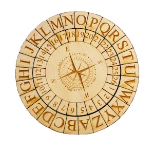 Puede incluir: Una rueda de cifrado redonda de madera con un diseño de rosa de los vientos en el centro. El anillo exterior presenta el alfabeto y números, mientras que el círculo interior muestra una rosa de los vientos. La madera es de color marrón claro.