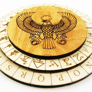 Op de afbeelding: Een rond houten divinatiebord met drie draaiende lagen. De bovenste laag heeft een lasergegraveerd beeld van een gevleugelde Egyptische god met een zonneschijf boven zijn hoofd. De middelste laag heeft een reeks Egyptische hiërogliefen. De onderste laag heeft een reeks letters.