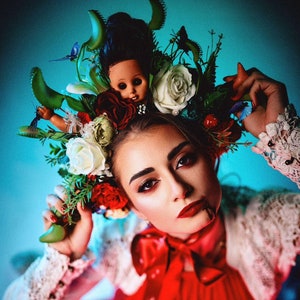 Puede incluir: Una mujer con un vestido rojo y una gran corona floral. La corona incluye una muñeca, flores y vegetación. La mujer lleva lápiz labial rojo y una mancha de sangre en la barbilla.