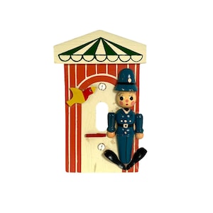 May include: A vintage wooden light switch cover featuring a whimsical design. The cover has a red and white striped background with a green and white awning. A blue-uniformed figure and a yellow bird are attached to the cover.