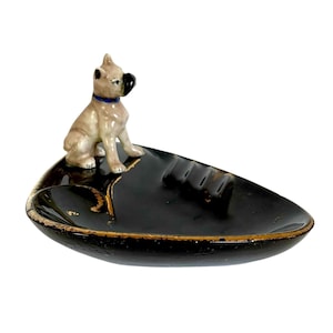 May include: Black ashtray with a gold trim and a ceramic dog figurine on top. The ashtray is shaped like a leaf and has grooves for cigarettes. The dog is tan with black ears and a blue collar.