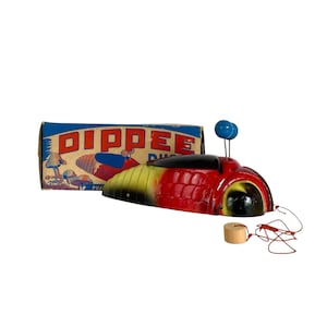 May include: A red and yellow toy with a black windshield and two blue antennae, accompanied by its original blue and white box labeled DIPPEE. A thin red string is attached to a small wooden cylinder, extending from the side of the toy.