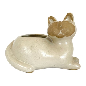 May include: A ceramic planter shaped like a white cat with brown markings. The cat is lying down with its head turned to the side. The planter has a small opening on the top for planting.