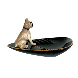 May include: A ceramic decorative dish with a black, leaf-shaped design. A tan-colored dog figurine with a blue collar sits on the dish. The dish has a gold-colored trim. The dog is looking to the left.