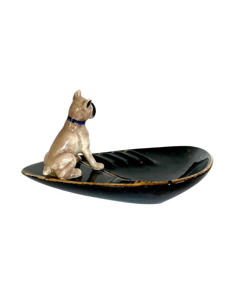 May include: A ceramic decorative dish with a black, leaf-shaped design. A tan-colored dog figurine with a blue collar sits on the dish. The dish has a gold-colored trim. The dog is looking to the left.